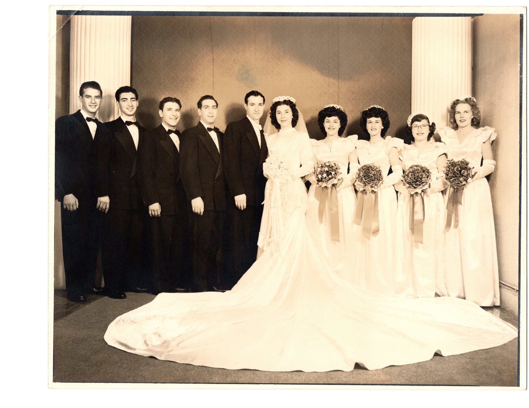 Formal wedding group portrait: five men in evening dress, the bride in a long train, and four bridesmaids with bouquets