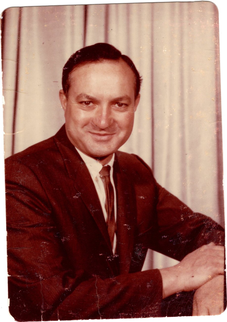 Colour portrait of Benny Enea in a dark jacket and tie, seated before a pale curtain