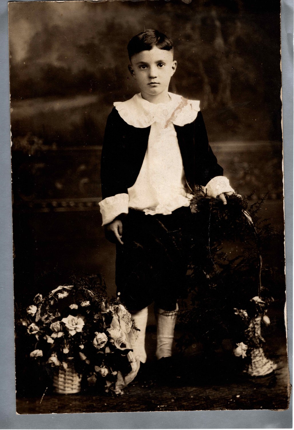 Studio portrait of a boy of about five in a velvet suit with a large white collar, standing beside baskets of roses