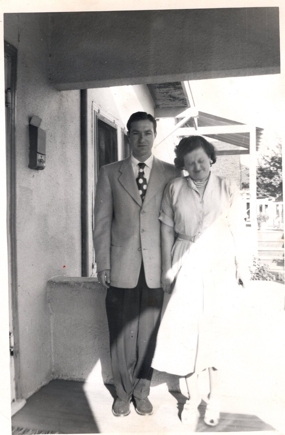 A young man in a light suit standing with his mother in a doorway, sunlight across the step
