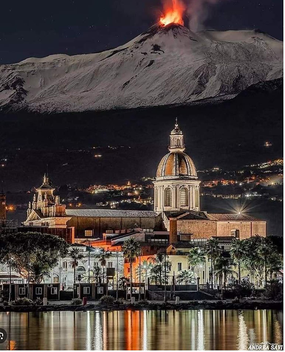 Nighttime view of Riposto, Sicily, with illuminated church and erupting Mount Etna beyond
