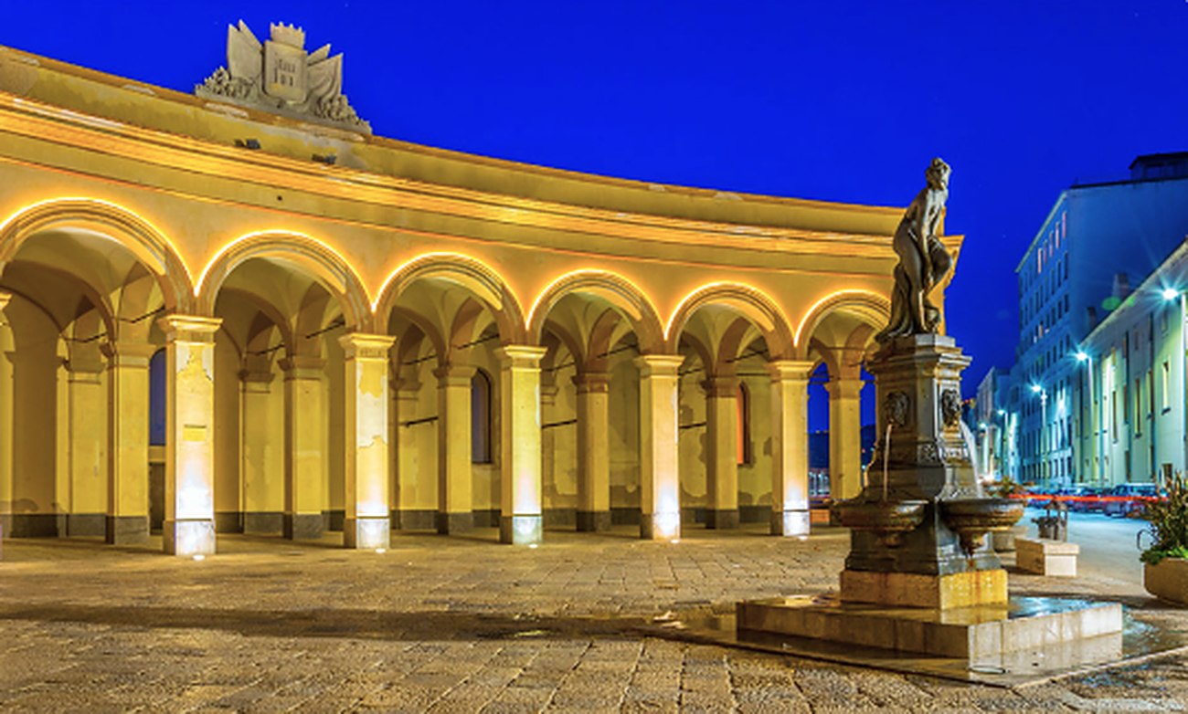 The curved colonnade of the Old Fish Market in Trapani, floodlit at dusk, with a statue on a pedestal