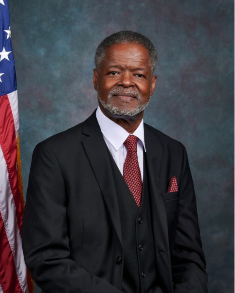 Official portrait of Federal Glover in a dark three-piece suit and red patterned tie, standing beside an American flag