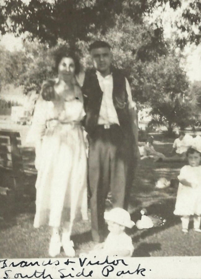 Frances Junta in a white dress standing beside Victor Giambona in a park, with a small child seated at their feet