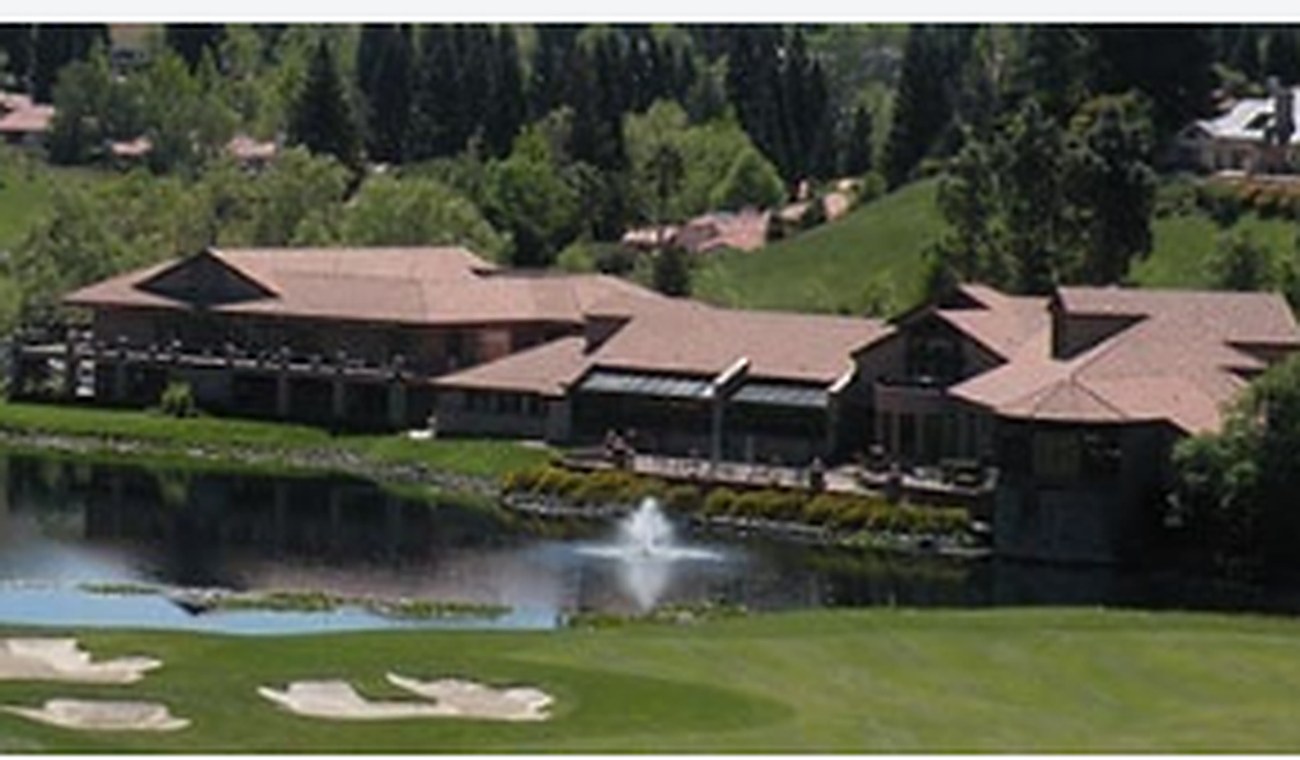 A low timber clubhouse beside a lake with a fountain, fairway and bunkers in the foreground