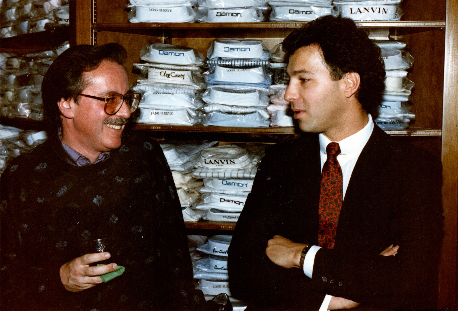 Two men standing among racks of suits and shirts inside a menswear store in the mid-1980s