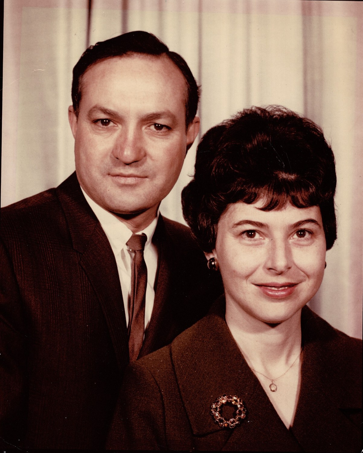 Colour studio portrait of a couple, the man in a dark suit and the woman in a brown jacket with a circular brooch