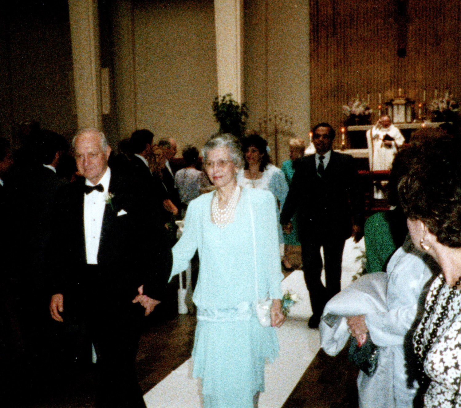 A couple walking up a church aisle after a wedding, the woman in a pale blue dress and pearls