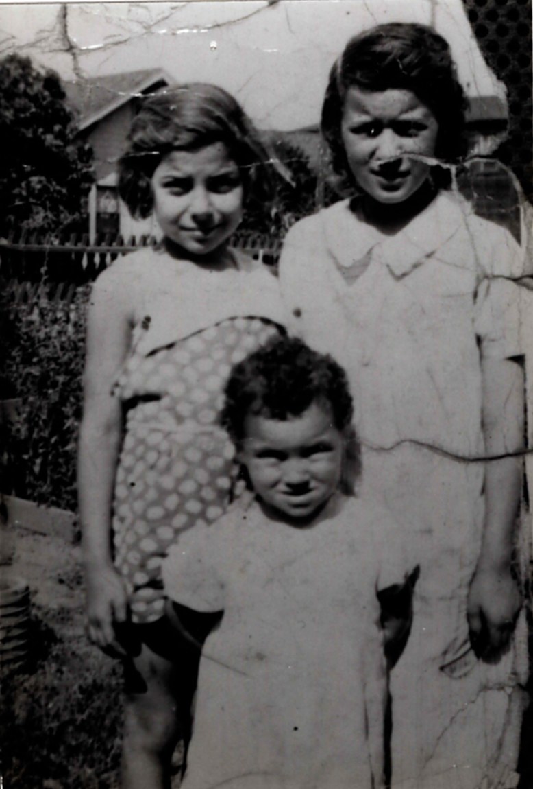 Three children standing in a backyard, two girls behind a smaller child, in front of a wooden fence and house