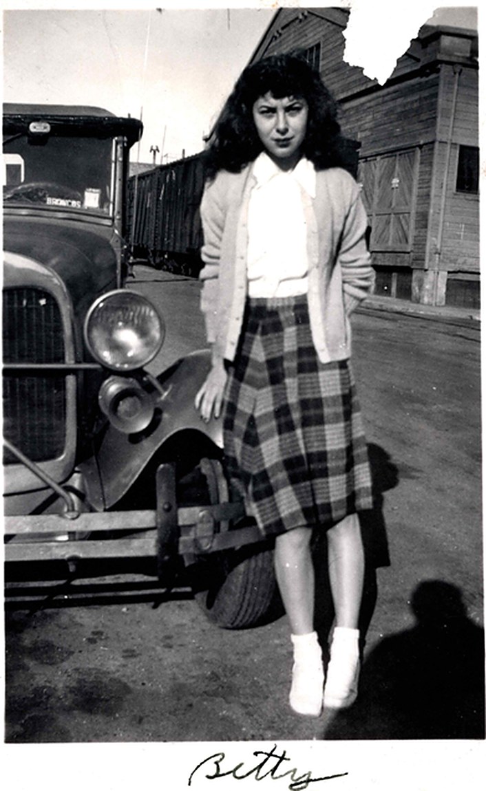 A teenage girl in a cardigan and plaid skirt standing beside the front fender of a 1930s automobile