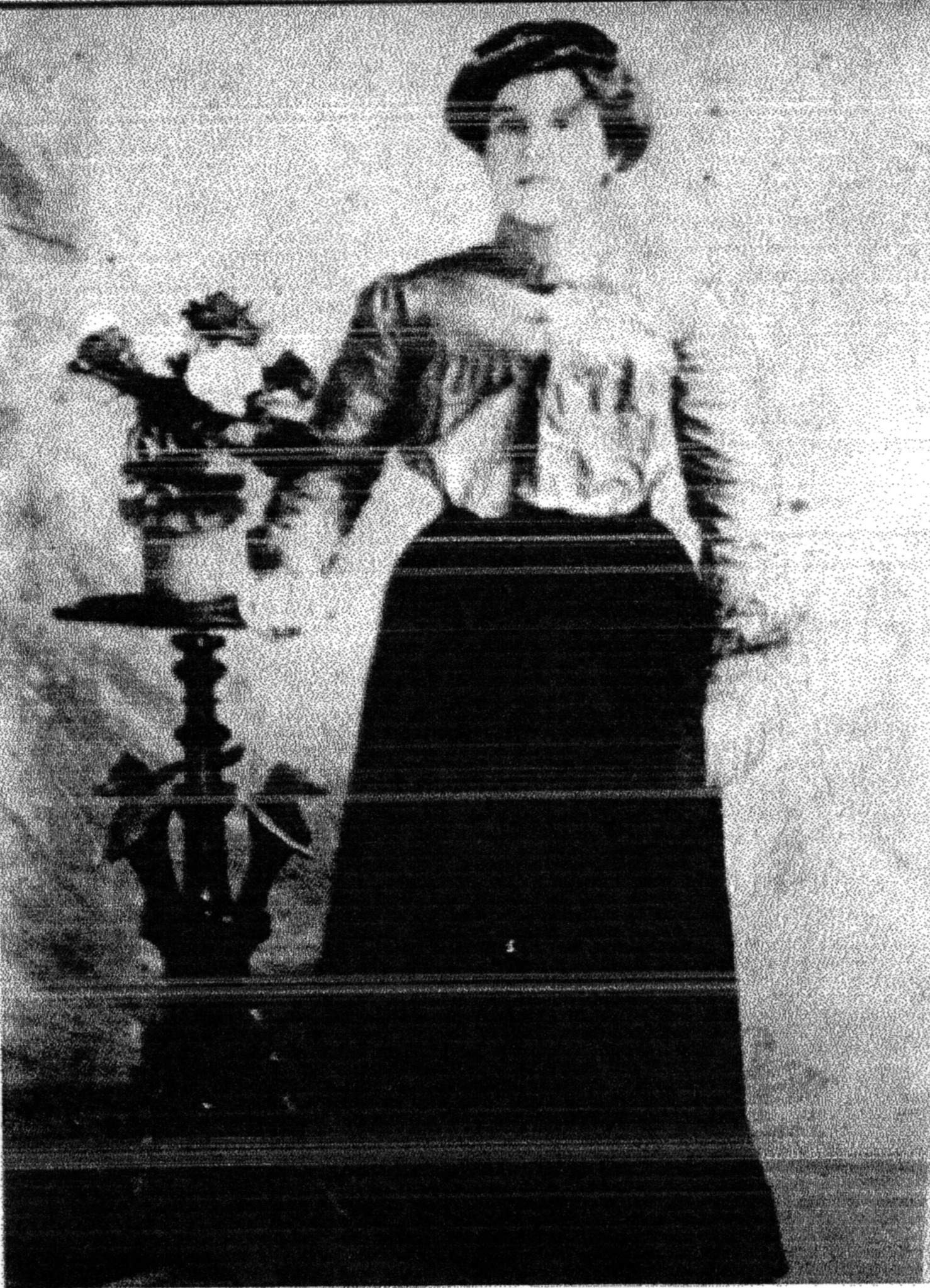 A young woman in a high-necked blouse and long dark skirt standing beside a pedestal table of flowers