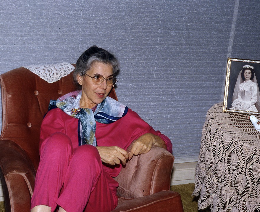 An older woman in a pink outfit and patterned scarf seated in an armchair, her framed wedding portrait on the lace-covered table beside her