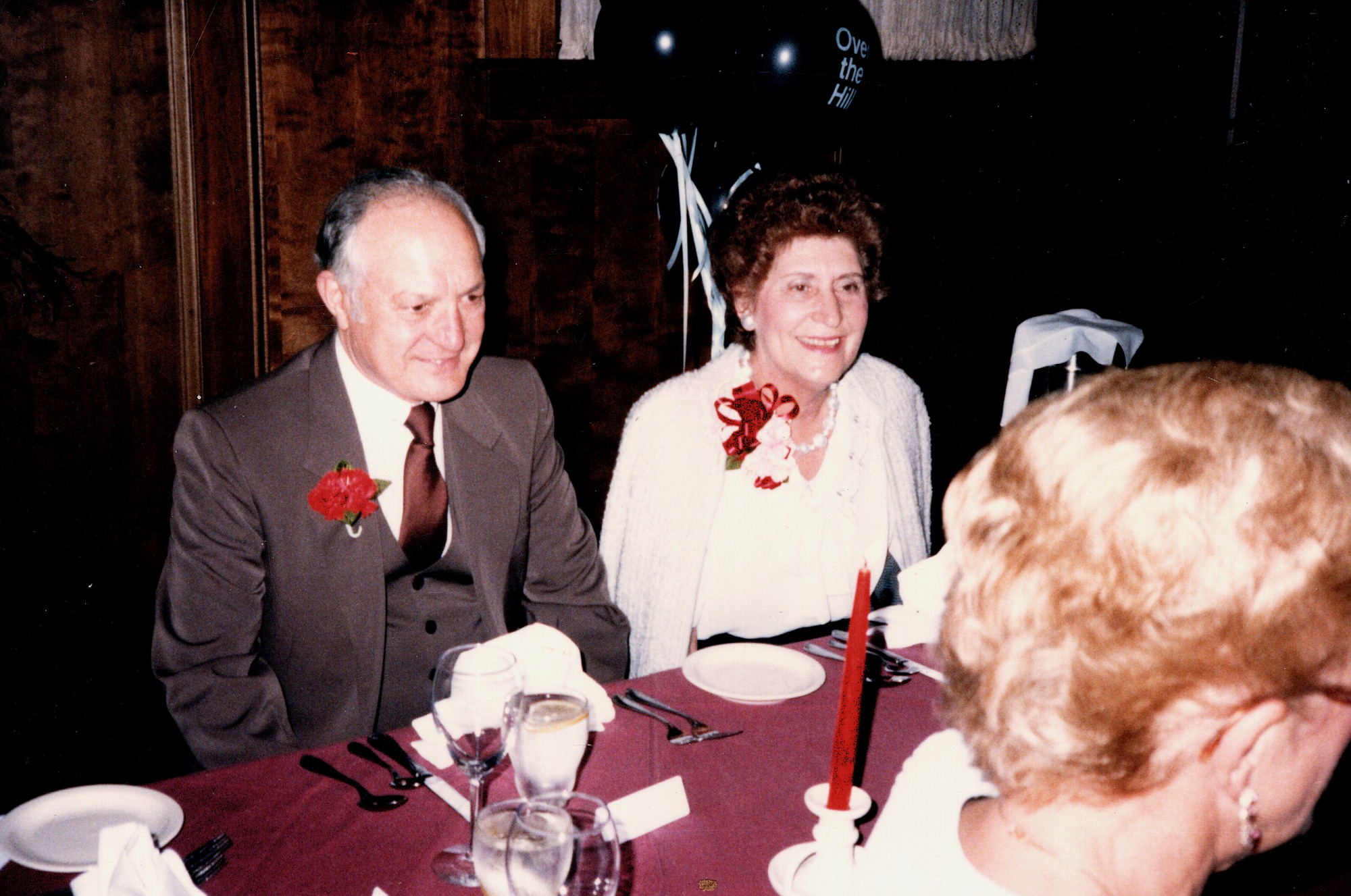 A man in a suit with a red carnation and a woman in a white jacket seated together at a banquet table