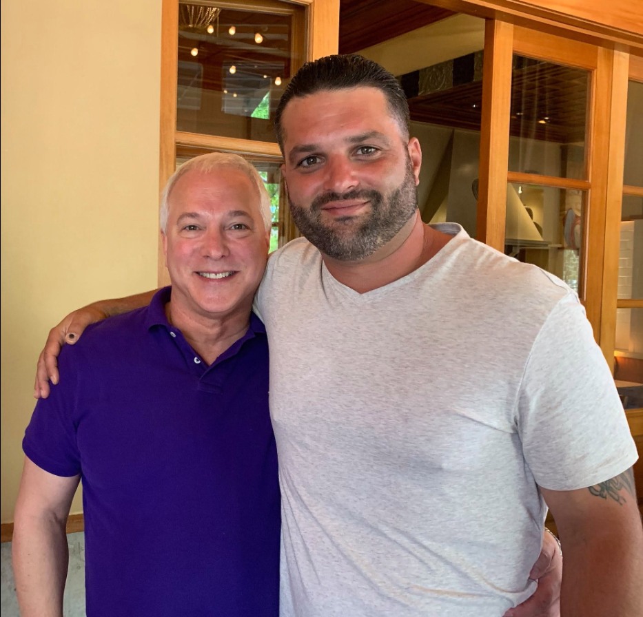 Two men standing arm in arm in a restaurant, one in a purple polo shirt and the other in a grey t-shirt