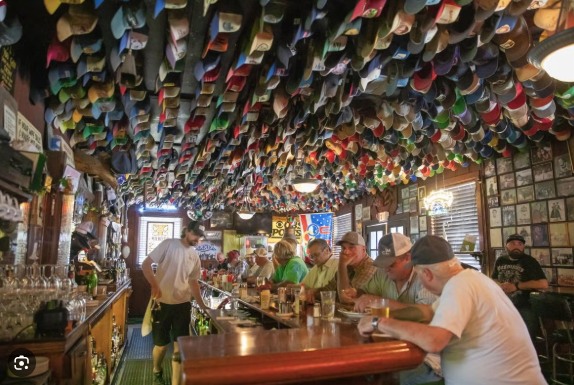 The interior of a bar, its ceiling covered in hundreds of baseball caps, with patrons seated along a long wooden counter