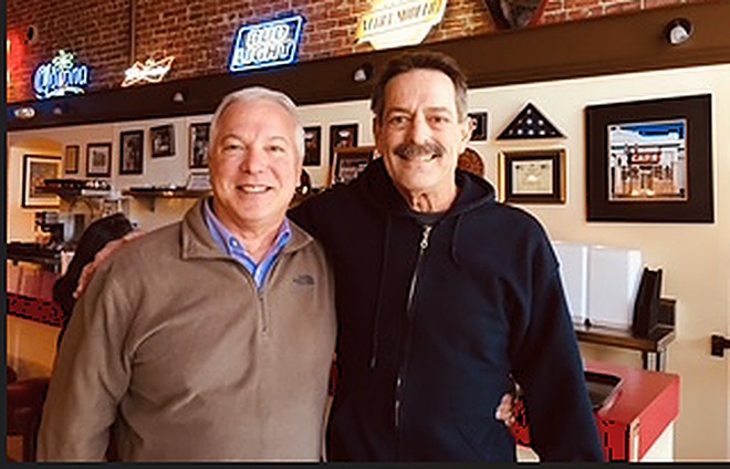 Two men standing side by side inside a restaurant, one with an arm around the other
