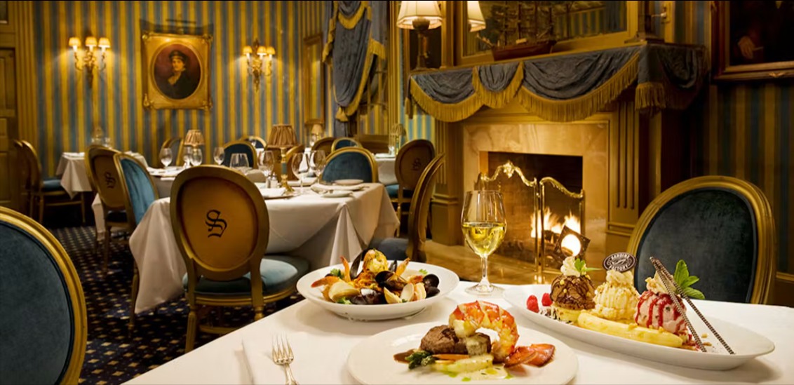 A formal dining room with striped walls, gold chairs and a lit fireplace, a table set with dishes in the foreground