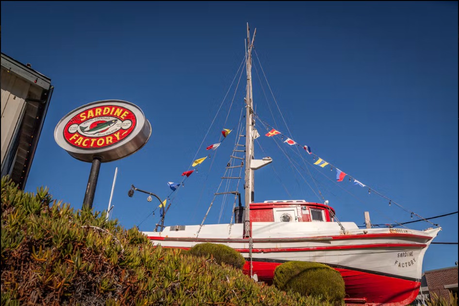 The Sardine Factory sign beside a red and white fishing boat mounted outside the restaurant under a clear blue sky
