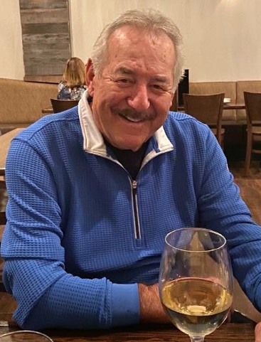 A smiling man in a blue quarter-zip sweater seated at a restaurant table with a glass of white wine