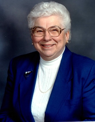 Studio portrait of an older woman with white hair and glasses in a blue jacket and pearls
