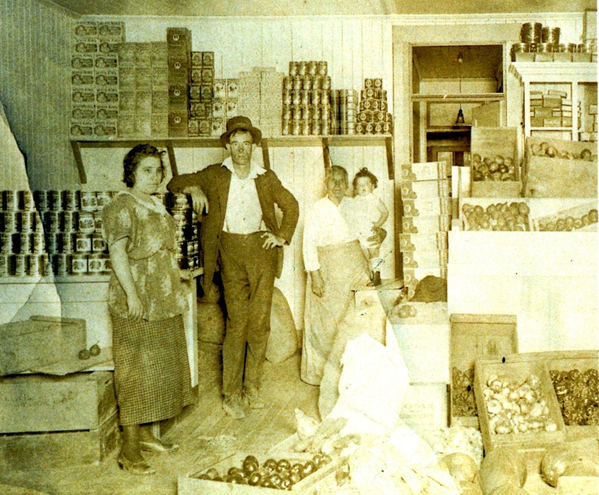 Maria Cardinale-Riccabuano standing in her market among stacked crates and shelves of canned goods, holding the infant Frances Junta