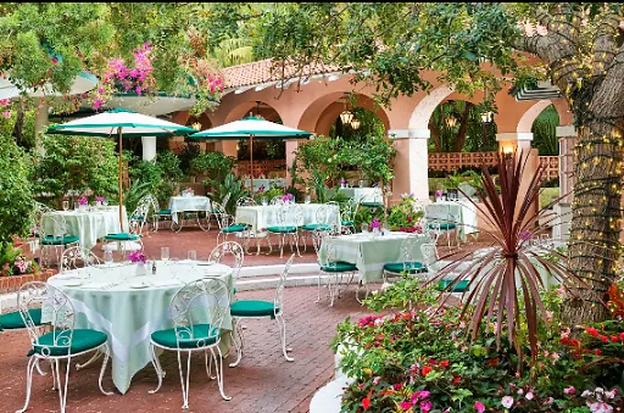 The Polo Lounge patio: white wrought-iron tables under green umbrellas, bougainvillea and pink stucco arches