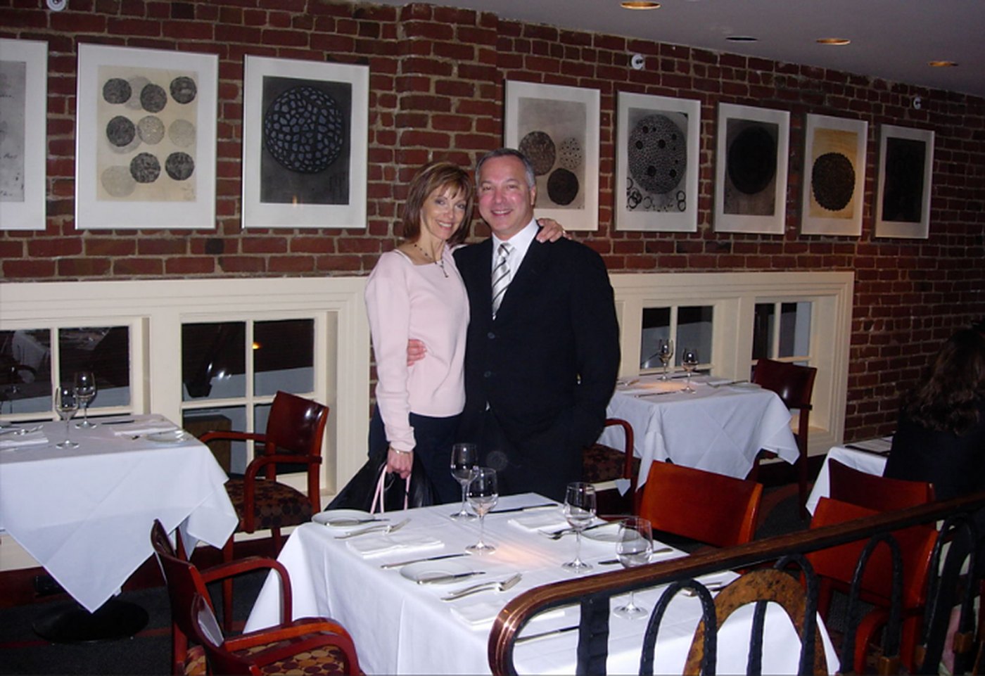 Tami and Charles Enea standing beside a set table in the dining room at Postrio, brick walls hung with framed prints