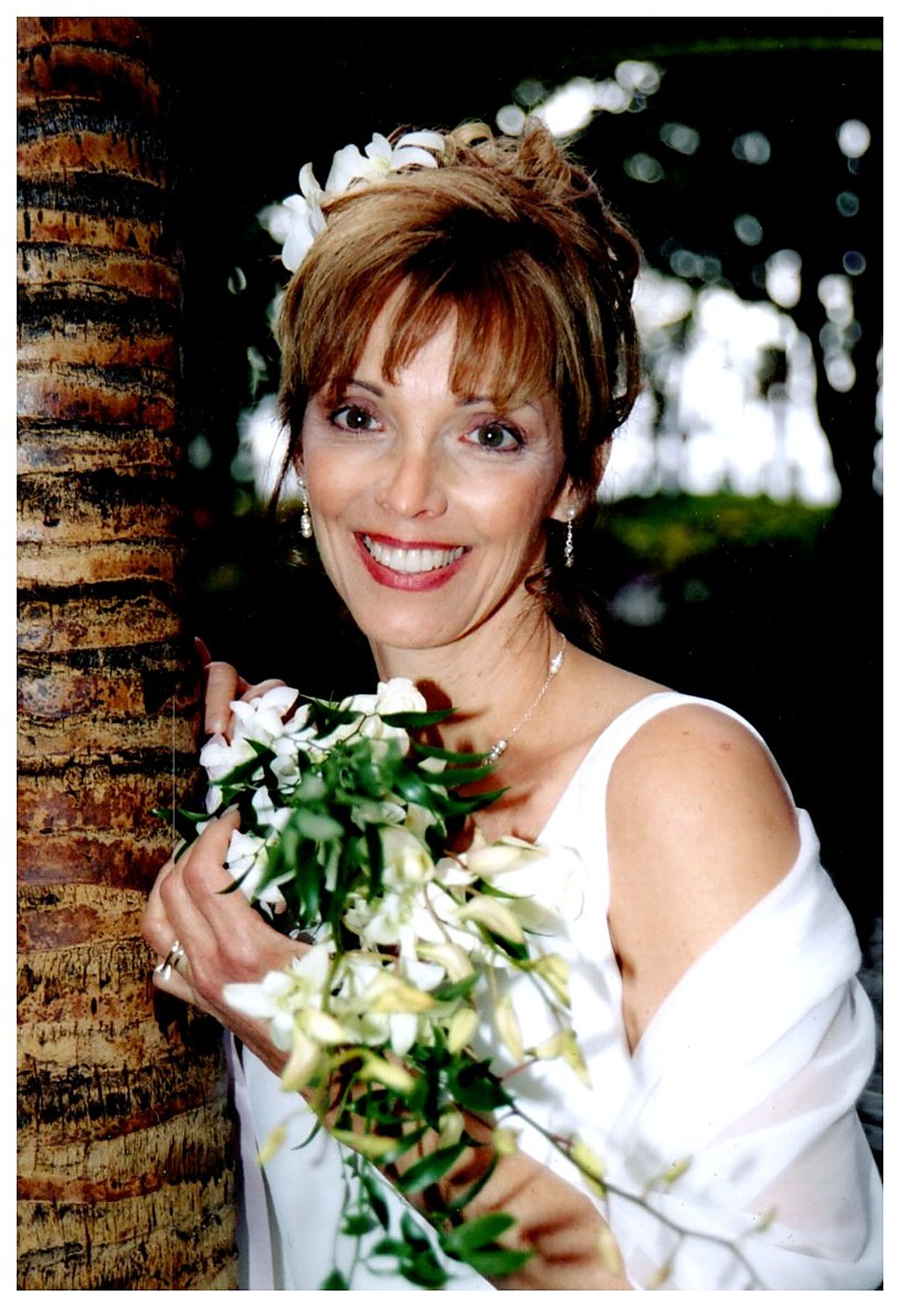Tami on her wedding day, holding a cascading white orchid bouquet beside a palm trunk